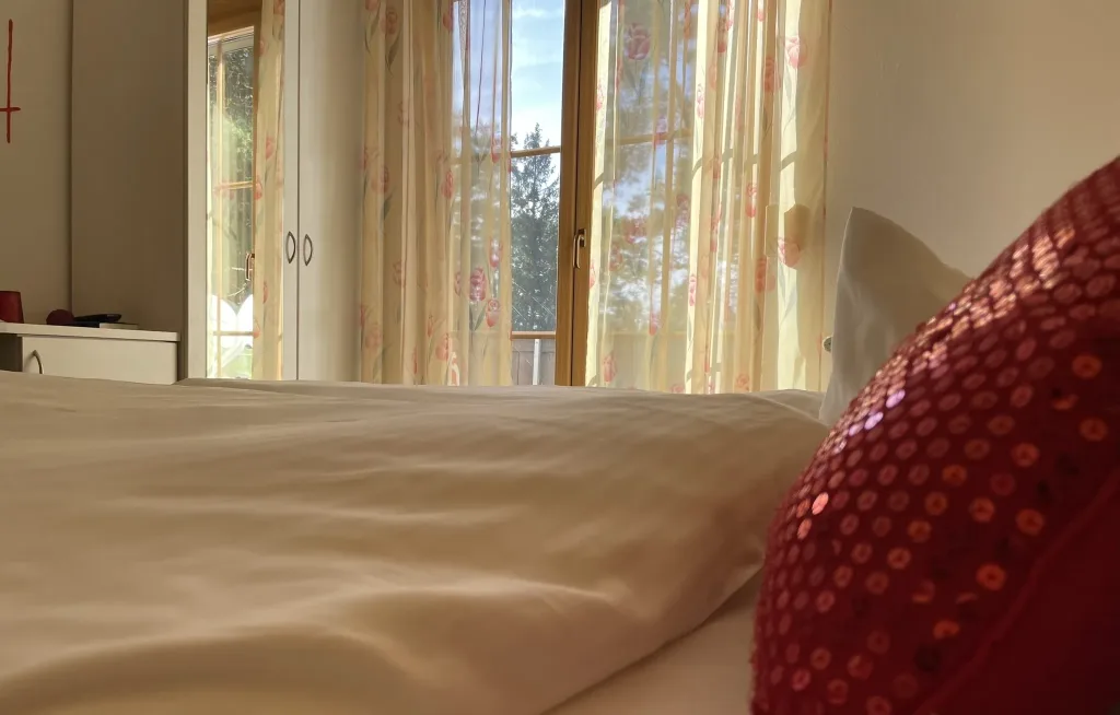 Hotel bed with a red cushion, behind it the balcony door with a view of the greenery
