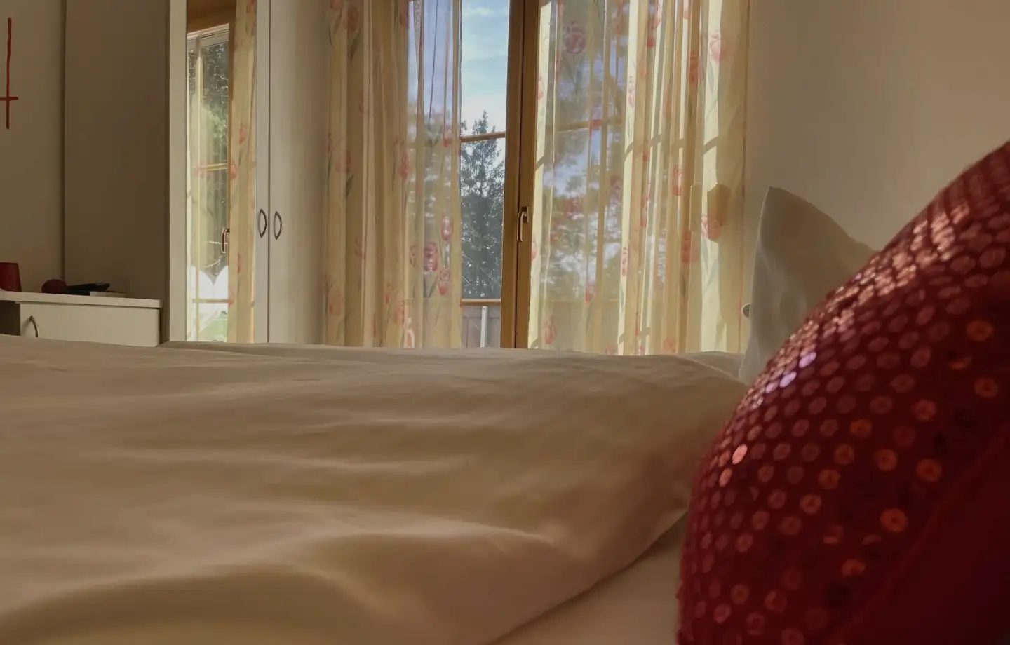 Hotel bed with a red cushion, behind it the balcony door with a view of the greenery